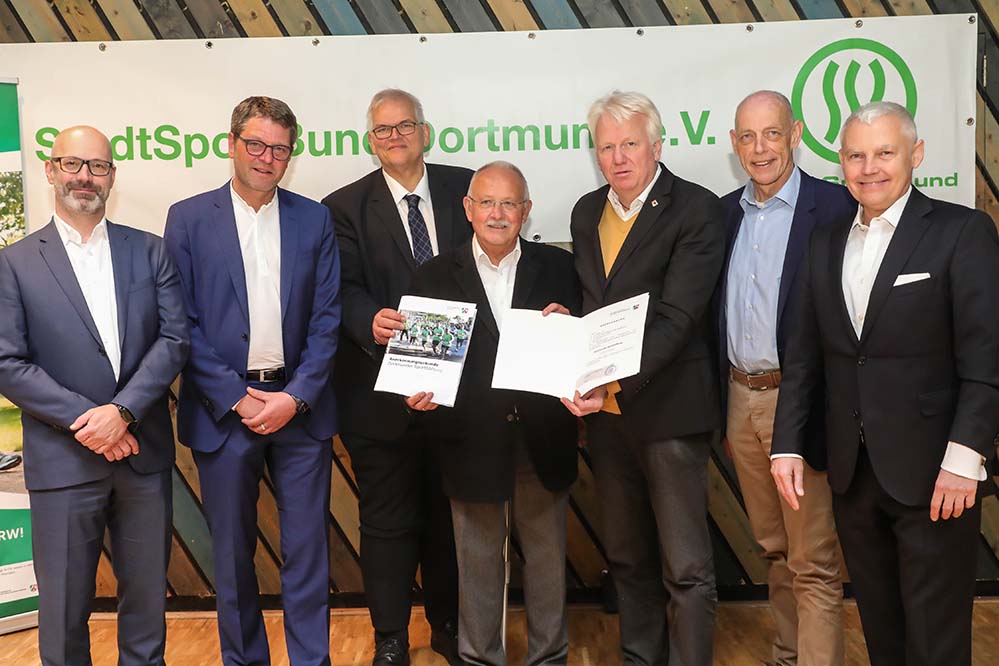 Anfang April 2019 richtete der StadtSportBund Dortmund e. V. seine 50. ordentliche Mitgliederversammlung in der Aula der CJD Jugenddorf-Christophorusschule aus. Zahlreiche Delegierte aus unterschiedlichen Dortmunder Sportvereinen und viele Ehrengäste waren der Einladung des SSB Dortmund gefolgt. Oberbürgermeister Ullrich Sierau sprach in seinem Grußwort dem organisierten Sport mit seinen zahlreichen ehrenamtlichen Mitarbeitern seinen Dank und die Anerkennung für die geleistete Arbeit aus. Der StadtSportBund, als Dachverband von über 500 Sportvereinen, mit mehr als 150.000 Mitgliedern, bildet in vielen Belangen das Bindeglied zwischen der Lokalpolitik, der städtischen Verwaltung und den Sportvereinen. Nicht zuletzt als Zeichen der Anerkennung für diese Arbeit wurde nicht nur die 20%ige Kürzung der Sportfördermittel aufgehoben, sondern eine Erhöhung um weitere 500.000,00 € im Haushaltsjahr 2019 verabschiedet. Damit stehen dem Sport in Dortmund im Jahr 2019 insgesamt 1,0 Millionen € zur Verfügung. Des Weiteren ging Herr Sierau auf die Sporthallensituation in der Stadt ein und berichtete über geplante Maßnahmen und Abstimmungsprozesse. Sehr zur Freude des Dortmunder Sports übergab der Regierungspräsident Hans-Josef Vogel die Stiftungsurkunde der „Dortmunder SportStiftung“ persönlich an den Vorstandsvorsitzenden des SSB und neuen Vorsitzenden der „Dortmunder SportStiftung“ Jörg Rüppel. Die vier in Dortmund ansässigen Unternehmen Sparkasse Dortmund, WILO SE, DSW21 und Adesso AG bilden als Gründungsstifter das finanzielle Fundament der zukunftsorientierten Leistungssportförderung durch die „Dortmunder SportStiftung“ und setzen mit ihrem Engagement insbesondere für den Leistungssport in dieser Stadt ein eindrucksvolles Zeichen. Geschäftsführer Mathias Grasediek berichtete über die geplante Neufassung der Sportförderrichtlinien. Gemeinsam mit den Sport- und Freizeitbetrieben wurde die dem Rat demnächst zur Abstimmung ausgearbeitete Vorlage abgestimmt. So sollen die Zuschüsse für die Vereine zur Förderung der Jugend- und der Übungsarbeit ebenso deutlich angehoben werden wie die Zuschüsse zur Anschaffung vereinseigener Grundsportgeräte. Zusätzlich sieht der Entwurf der neuen Richtlinien vor, lizenzierte Sportvereinsmanager deutlich höher zu unterstützen sowie Sportvereinen Zuschüsse zur Ausbildung qualifizierte Vereinsmanager zu ermöglichen. Diese Vereinsmanageroffensive liegt dem SSB besonders am Herzen, da qualifizierte Kräfte im Bereich der Vereinsführung die Zukunftsfähigkeit der Vereine nachhaltig stärken und unterstützen werden. In einer Umfrage richtete sich der SSB im vergangenen Jahr an seine Mitglieder, um herauszufinden, was die Ehrenamtlichen antreibt sich im Sport zu engagieren und wo in den Vereinen der Schuh drückt. Weiter wurde abgefragt wie die Sportvereine mit dem SSB und dritten Partnern rund um den Sport kooperieren und wo die Sportvereine zukünftig die Arbeitsschwerpunkte des SSB sehen. In einem kurzen Überblick stellte Nadine Euler einige Ergebnisse vor. Einen konkreten Arbeitsauftrag nahm der Vorstand direkt aus der Befragung mit: 68 % der Teilnehmenden wünschen sich ein Führungskräftetreffen, auf dem sich die Vorstände der Sportvereine untereinander austauschen können. In Anerkennung seiner langjährigen und verdienstvollen Tätigkeit für den Dortmunder Sport wurde Jürgen Grondziewski mit der Goldenen Ehrennadel des SSB ausgezeichnet. Der SSB und seine Sportjugend selbst erhielten ebenfalls eine bedeutsame Anerkennung. Als erster Sportbund in NRW wurde er in das Qualitätsbündnis gegen sexualisierte Gewalt im Sport in NRW aufgenommen. Dieses Bündnis hat maßgeschneiderte Qualitätsstandards zur Prävention und Intervention entwickelt. Eine seiner wichtigsten Ziele ist es, die Sportvereine dabei zu unterstützen, die Prävention gegen sexualisierte Gewalt in ihrem Verein zu verankern und eine Kultur der Achtsamkeit zu entwickeln. Dr. Birgit Palzkill (unabhängige Beauftragte zum Schutz vor sexualisierter Gewalt im Sport des Landessportbund NRW) ließ es sich nicht nehmen, diese Urkunde persönlich zu überreichen. Bildzeile v. l. n. r.: Stiftungsurkunde Dortmunder SportStiftung überreicht: A. Prenneis (Adesso AG), K. Hoffmann (WILO SE), H.-J. Vogel (Regierungspräsident), J. Rüppel (Vors. SSB), U. Sierau (OB), K. Steenweg (Sparkasse), Dr. Puplick (Rechtsanwalt)