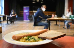 Das Foto zeigt die neue Essensausgabe im großen Saal des Wichernhauses. 20 wohnungslose Menschen können hier ab sofort dreimal pro Woche gleichzeitig eine warme Mahlzeit zu sich nehmen, die täglich von der Diakonie frisch zubereitet wird. Nach jeder Mahlzeit wird der Platz gereinigt und ein neuer Gast hereingeführt. Dieses System ermöglicht wohnungslosen Menschen zum ersten Mal seit März, eine warme Speise vor Ort einzunehmen und hat das bisherige System mit den Lunchpaketen im Außenbereich des Wichernhauses abgelöst. Foto: Diakonie