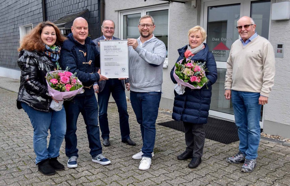 Gleich ein doppeltes Jubiläum galt es jetzt beim Dachdeckerbetrieb Klotzbach GmbH in Dortmund-Derne zu feiern. Nicht nur, dass der Betrieb in diesem Jahr sein 70. Gründungsjubiläum begeht, auch Juniorchef Denis Struwe konnte jetzt auf sein 25-jähriges Jubiläum als Dachdeckermeister zurückblicken. Aus diesem Anlass besuchten der Obermeister der Dachdecker-Innung Dortmund und Lünen, Dirk Sindermann, und Innungsgeschäftsführer Ludgerus Niklas das Traditionsunternehmen und überreichten den Silbernen Meisterbrief an den 49-jährigen Jubilar. „Es ist mir eine ganz besondere Ehre, heute in einem Betrieb zu sein, der in unserem Handwerk schon seit so vielen Jahren erfolgreich arbeitet“, so Obermeister Dirk Sindermann in seiner Laudatio. „Mein besonderer Glückwunsch, den ich auch im Namen aller Mitgliedsbetriebe der Innung überbringe, gilt jedoch Denis Struwe, der als Dachdeckermeister nun schon seit 25 Jahren in dritter Generation diese Tradition fortführt.” Seit drei Generationen in Derne 1951 hatte der Dachdeckermeister Otto Klotzbach seinen Betrieb „Das Dach Klotzbach GmbH“ in Dortmund-Derne gegründet. Mit nur einem Angestellten und einer Handkarre begann er, eine Existenz aufzubauen. Qualität und Zuverlässigkeit ließen das Unternehmen stetig wachsen. Manfred Struwe, der heutige Seniorchef und Ehrenobermeister der Dachdecker-Innung Dortmund und Lünen, heiratete in den Betrieb ein und übernahm ihn 1981. Sein ältester Sohn Denis Struwe lernte sehr früh über den Großvater und Vater das Handwerk kennen. Selbst vom Beruf des Dachdeckers begeistert, absolvierte er im Betrieb eine Lehre und machte seine Ausbildung zum Meister am Bildungszentrum des westfälischen Dachdeckerhandwerks im sauerländischen Eslohe. 1996 schloss er diese mit der erfolgreichen Prüfung vor der Handwerkskammer Arnsberg ab. Kurz danach wechselte er in die Geschäftsführung des Betriebs und wurde Mitgesellschafter. Seniorchef Manfred Struwe, der mittlerweile 74 Jahre alt ist, freut sich darüber, dass das Unternehmen bei seinem Sohn in guten Händen ist, und unterstützt ihn bei Bedarf. Breites Portfolio und große Fachkenntnis Das Dach Klotzbach GmbH hat heute 13 Beschäftigte, arbeitet für private und öffentliche Auftraggeber mit einem breiten Portfolio an fachlich hochwertigen Leistungen. Sie reichen von klassischen Dachdeckerarbeiten an Flach- und Steildächern inklusive Reparatur und Wartung über die Bauklempnerei bis zum Einbau von Dachfenstern und der Installation von Solaranlagen und Dachbegrünungen. Denis Struwe lenkte den Betrieb schon früh in die Digitalisierung, machte 2002 seinen „Manager im Dachdeckerhandwerk” beim Berufsbildungswerk des Deutschen Dachdeckerhandwerks und der European Business School und beschreitet heute mit dem Betrieb konsequent dem Weg ins digitale „Handwerk 4.0”. Die Tradition wird dabei allerdings nicht vergessen. Schon seit seiner Gründung im Jahr 1951 ist der Betrieb Innungsmitglied. Seniorchef Manfred Struwe war lange Jahre Obermeister der Dachdecker-Innung Dortmund und Lünen und Vorsitzender des Innungsverbandes des Dachdeckerhandwerks Westfalen. Heute ist er Ehrenobermeister der Innung. Jubilar Denis Struwe ist ebenfalls in der Innung aktiv. Er ist Vorsitzender der Prüfungskommission und damit für das Prüfungswesen verantwortlich. Bildzeile: Glückwunsch zum Silbernen Meisterbrief mit (v.l.)Tiziana Di Matteo-Struwe, Jubilar Denis Struwe, Innungsgeschäftsführer Ludgerus Niklas, Obermeister der Dachdecker-Innung Dortmund und Lünen Dirk Sindermann, Brigitte Struwe und Manfred Struwe. Foto: Dachdecker-Innung Dortmund und Lünen.