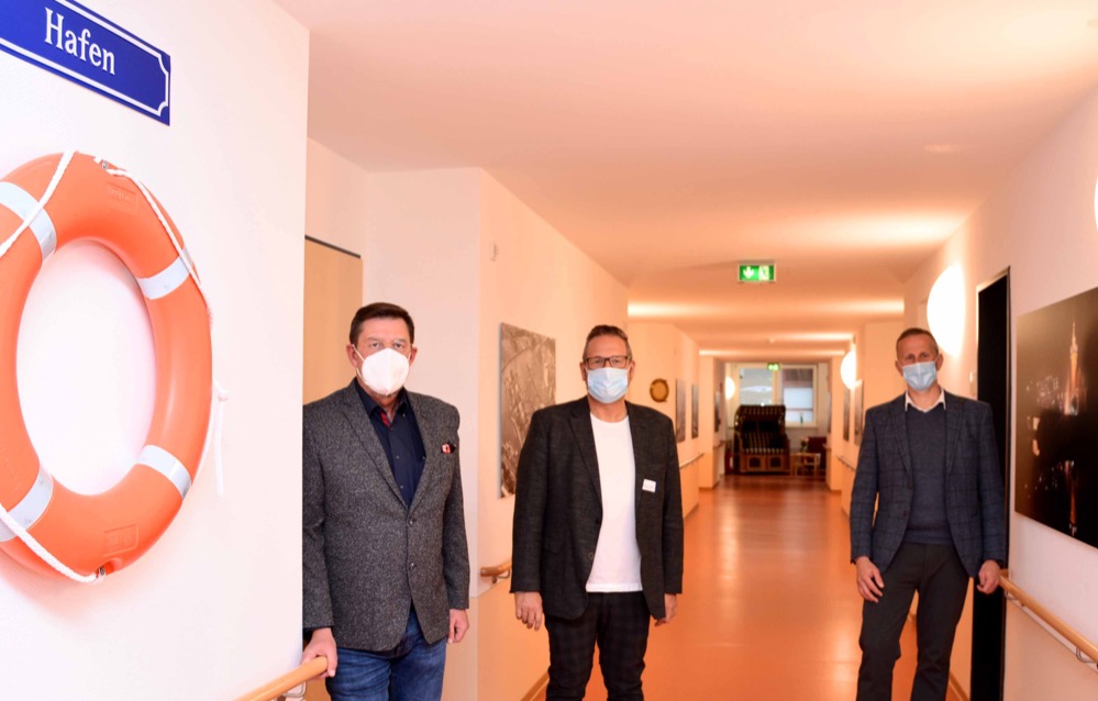 Der Dortmunder Hafen erstreckt sich neuerdings bis zum Nordmarkt: Im Evangelischen Altenzentrum „Der Gute Hirte“ hat die Diakonische Altenhilfe mit Unterstützung der Dortmunder Hafen AG in einem kompletten Stockwerk des Hauses an der Spohrstraße eine durchgängig maritime Gestaltungslinie verwirklicht. Historische Bilder und aktuelle Fotos lassen Dortmunds traditionsreiche Logistikdrehscheibe erlebbar werden. Großformatige Fotos, Bullauge, Rettungsring, Strandkorb und mehr: Jeder der Bewohner und jede Bewohnerin kann sich künftig wie beim Flanieren entlang einer Kaikante fühlen, was unter den Menschen des Hauses sehr gut ankommt und die Wohnzufriedenheit nochmals steigert. Um das Ergebnis der Kooperation zu feiern und die neue Einrichtung des Hauses vorzustellen, begrüßte am Montag, 8. November 2021, Einrichtungsleiter Stephan Eull den Vorstand der Dortmunder Hafen AG, Uwe Büscher, sowie den Geschäftsführer der Diakonischen Altenhilfe Dortmund und Lünen gGmbH, Jens Fritsch. Stephan Eull, Leiter des Guten Hirten, will nun nach und nach auch die weiteren drei Etagen mit Nordstadt-Örtlichkeiten in Verbindung bringen. Etage zwei nimmt sich den Fredenbaum vor, die dritte den Nordmarkt und die vierte den Borsigplatz. „Uns geht es darum, unseren Bewohnern im Haus zusätzliche Orientierungshilfen zu geben und das Wohlbefinden zu steigern“, unterstreicht er. Das ist gelungen. Historische Bilder und aktuelle Fotos kennzeichnen den Hafen als modernes Wirtschaftsareal mit langer Geschichte. Vom Foto links grüßen die Güterschiffe „Delta“ und „Michaela“, auf dem Foto gegenüber türmen sich unterschiedliche Schüttgüter und veranschaulichen die Leistungsfähigkeit der Logistikdrehscheibe. Und wenn man so will: Wer hochbetagt auf der Etage wohnt, hat einen Großteil davon zumindest mitbekommen. Das ist das Nordstadtgefühl, das Eull transportieren möchte. Ein Foto des Alten Hafenamtes, dessen Silhouette sich nachts im Wasser spiegelt, gibt es auch. Und die Schiffsglocke aus Messing, die ebenfalls auf dem Flur an der Wand hängt, wird ab und zu von den Bewohnern geläutet: Heimatkunde im Vorübergehen. Wer in seinem privaten Fundus noch etwas hat, was thematisch auf die vier Etagen passt, ist herzlich eingeladen, sich im Altenzentrum „Der gute Hirte“ zu melden. Tel. (0231) 72 54 710. Foto: Uwe Büscher, Vorstand der Dortmunder Hafen AG, Einrichtungsleiter Stephan Eull und Jens Fritsch, Geschäftsführer der Diakonischen Altenhilfe Dortmund und Lünen gGmbH im Ev. Altenzentrum „Der Gute Hirte“ am Nordmarkt.