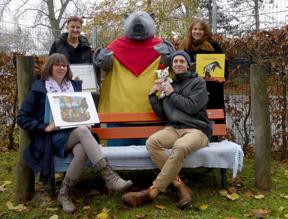 Das Netzwerk INFamilie Libellenquartier weihte am 19.11.2021 im Rahmen einer Vorleseaktion mit Besuch des Lesenashorns Plappermaul die „Vorlesebank“ für das Quartier ein. Zu finden ist die Vorlesebank in einer ruhigen Ecke des Außengeländes des AWO Familienzentrums / Kita Burgholzstraße und stellt einen Ort der Möglichkeiten für verschiedene Sprachanlässe im pädagogischen Alltag der Kindertageseinrichtungen des Quartiers sowie für die Arbeit mit engagierten Menschen aus der Nachbarschaft dar. Schüler*innen aus der benachbarten Libellengrundschule können hier den jüngeren Kindern beim Lesen von Texten ein Stück weit Vorbild sein und so ebenfalls ihre eigenen sprachlichen Fertigkeiten trainieren. Auch die älteren Bewohner*innen des Quartiers dürfen auf diese Weise in Zukunft an der Gestaltung des Libellenquartiers mitwirken: Als Lesepaten können sie sich in dem Zusammenhang mit den Kindern unterhalten, Bücher vorlesen und vielleicht auch die ein oder andere Geschichte aus ihrer eigenen Kindheit erzählen. Denn gerade solche Geschichten sind für die Kinder meist neu und dementsprechend sehr interessant. Die Möglichkeiten der Partizipation sowie der Herausforderung den Kindern den Spaß am Lesen und Zuhören zu vermitteln wird mit dieser Bank so ein Stück weit Folge geleistet. Alle am Projekt beteiligten Personen waren entsprechend guter Dinge, dass die Vorlesebank den gewünschten Beitrag für das Libellenquartier erzielen wird. Pia Tenberge, Leitung des AWO Familienzentrums / Kita Burgholzstraße sagte dazu: „Wir sind froh die Vorlesebank zu haben, damit wir insbesondere die Menschen aus dem Quartier teilhaben lassen können“. Sarah Herbst, Sozialarbeiterin der Präventionsfachstelle des Dortmunder Jugendamtes und Koordinatorin des Netzwerks INFamilie Libellenquartier, ist vollauf zufrieden mit dem Projekt: „Ich freue mich, dass wir mit diesem Förderangebot den Kindern im Quartier die Freude am Lesen in attraktiver Atmosphäre näher bringen können“ und auch Christiane Mika, Rektorin der Libellengrundschule, findet es vor allem schön, „dass hier besonders Kinder mit Kindern arbeiten und so voneinander lernen können“.
