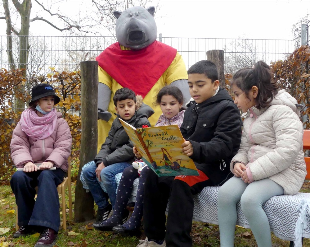Das Netzwerk INFamilie Libellenquartier weihte am 19.11.2021 im Rahmen einer Vorleseaktion mit Besuch des Lesenashorns Plappermaul die „Vorlesebank“ für das Quartier ein. Zu finden ist die Vorlesebank in einer ruhigen Ecke des Außengeländes des AWO Familienzentrums / Kita Burgholzstraße und stellt einen Ort der Möglichkeiten für verschiedene Sprachanlässe im pädagogischen Alltag der Kindertageseinrichtungen des Quartiers sowie für die Arbeit mit engagierten Menschen aus der Nachbarschaft dar. Schüler*innen aus der benachbarten Libellengrundschule können hier den jüngeren Kindern beim Lesen von Texten ein Stück weit Vorbild sein und so ebenfalls ihre eigenen sprachlichen Fertigkeiten trainieren. Auch die älteren Bewohner*innen des Quartiers dürfen auf diese Weise in Zukunft an der Gestaltung des Libellenquartiers mitwirken: Als Lesepaten können sie sich in dem Zusammenhang mit den Kindern unterhalten, Bücher vorlesen und vielleicht auch die ein oder andere Geschichte aus ihrer eigenen Kindheit erzählen. Denn gerade solche Geschichten sind für die Kinder meist neu und dementsprechend sehr interessant. Die Möglichkeiten der Partizipation sowie der Herausforderung den Kindern den Spaß am Lesen und Zuhören zu vermitteln wird mit dieser Bank so ein Stück weit Folge geleistet. Alle am Projekt beteiligten Personen waren entsprechend guter Dinge, dass die Vorlesebank den gewünschten Beitrag für das Libellenquartier erzielen wird. Pia Tenberge, Leitung des AWO Familienzentrums / Kita Burgholzstraße sagte dazu: „Wir sind froh die Vorlesebank zu haben, damit wir insbesondere die Menschen aus dem Quartier teilhaben lassen können“. Sarah Herbst, Sozialarbeiterin der Präventionsfachstelle des Dortmunder Jugendamtes und Koordinatorin des Netzwerks INFamilie Libellenquartier, ist vollauf zufrieden mit dem Projekt: „Ich freue mich, dass wir mit diesem Förderangebot den Kindern im Quartier die Freude am Lesen in attraktiver Atmosphäre näher bringen können“ und auch Christiane Mika, Rektorin der Libellengrundschule, findet es vor allem schön, „dass hier besonders Kinder mit Kindern arbeiten und so voneinander lernen können“.