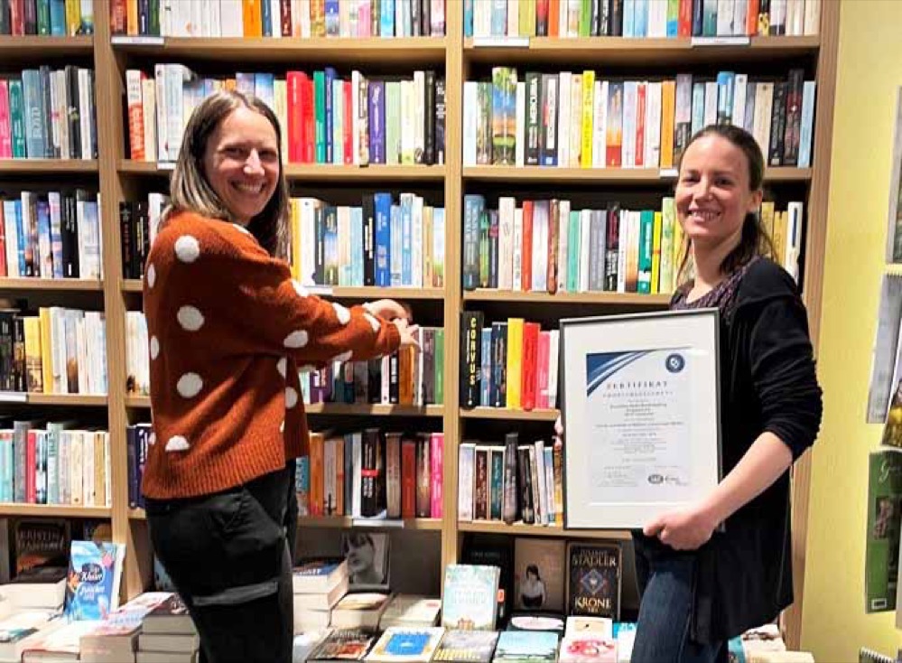 Nachhaltiges Wirtschaften ist gut für die Umwelt – und kann ein echter Vorteil bei Ausschreibungen sein. Ein Beispiel dafür ist die Dortmunder Bonifatius-Buchhandlung am Propsteihof. Die hat bereits im September 2020 das Zertifikat Umweltmanagement nach der DIN-Norm ISO 14001 erhalten. Jetzt wurde überprüft, ob die Buchhandlung die Vorgaben noch immer einhält. „Wir wurden erneut zertifiziert und damit unsere Arbeit bestätigt“, erklärt Eva Schulz, stellvertretende Filialleiterin in der Dortmunder Buchhandlung. Ganz praktisch bedeutet das für den Facheinzelhändler, der zur Paderborner Bonifatius GmbH gehört, vor allem Emissionen zu senken. Ware wird verstärkt zu Fuß oder per Fahrradkurier ausgeliefert, das eigene Fahrzeug wird für 2021 durch Ausgleichszahlungen klimaneutral gestellt. Zudem setzt die Buchhandlung bei Verpackungen auf Kartonagen, die zu 100 Prozent aus recyceltem Material bestehen. Und auch Energiefresser wie Neonröhren wurden ausgetauscht. „Wir haben den gesamten Workflow der Buchhandlung den Zertifizierungskriterien unterworfen und einen Umweltmanager hinzugezogen“, betont Rolf Pitsch, Geschäftsführer der Bonifatius GmbH. Dieser Aufwand lohnt sich nicht nur im Hinblick auf mehr Umweltschutz. Denn die Buchhandlung nimmt bundesweit an Ausschreibungen teil, wenn beispielsweise Städte Lieferanten für Schulbücher suchen. Kommunen und Bildungseinrichtungen erwarten dabei zunehmend die Einhaltung von Umweltstandards. Hier ist das Zertifikat ein großer Vorteil für den Fachhandel vor Ort, denn es wird bereits in den ersten Ausschreibungen explizit erwähnt. Zum Konzept gehört auch, dass die Bonifatius GmbH ihre eigenen Zulieferer mit ins Boot nehmen möchte. Dazu gehören auch die Buchgroßhändler, die über Nacht die Kundenbestellungen vom Vortag nach Dortmund bringen, damit sie dort vom Kunden persönlich abgeholt oder mit dem Fahrrad ausgeliefert werden können. Die Dortmund Bonifatius Buchhandlung ist eine von drei Buchhandlungen der Bonifatius GmbH, ein Medienunternehmen mit Sitz in Paderborn. Die Buchhandlung besteht seit mehr als 30 Jahren in der Innenstadt. Das Paderborner Unternehmen mit rund 160 Mitarbeitenden wurde 1869 als eigenständiges Unternehmen mit Druckerei, Buchhandlung und Buchverlag sowie katholischen Zeitschriften gegründet.