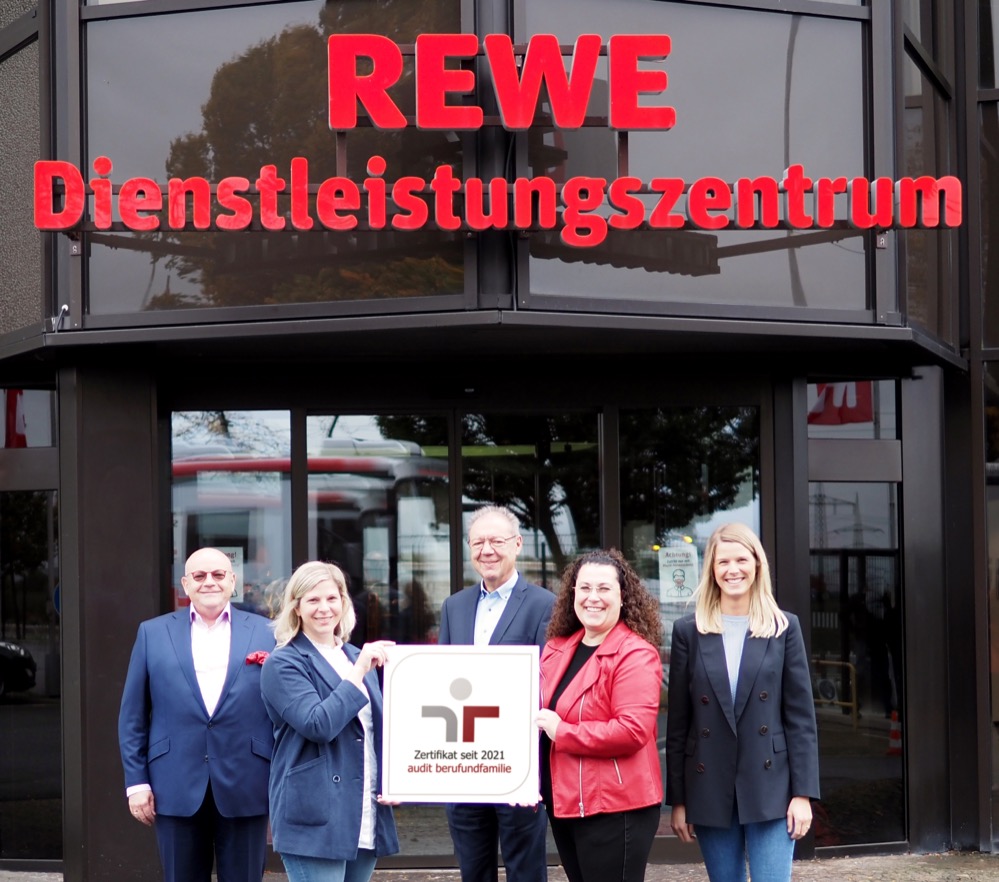 Die Vereinbarkeit von Beruf und Familie ist in der Arbeitswelt längst kein Randthema mehr. In Zeiten des Fachkräftemangels kommt der Work-Life-Balance eine starke Bedeutung zu. Im Dienstleistungszentrum der REWE Dortmund mit Verwaltung, Logistik und Produktion hat Familienfreundlichkeit schon eine lange Tradition. Jetzt ist das Großhandelsunternehmen mit dem Vereinbarkeitszertifikat der berufundfamilie Service GmbH ausgezeichnet worden und kann diese Philosophie nun auch nach außen zeigen. Das Zertifikat, das als Qualitätssiegel für eine betriebliche Vereinbarkeitspolitik gilt, wird von einem Kuratorium vergeben. Voraussetzung ist das erfolgreiche Durchlaufen des dazugehörigen Auditierungsprozesses. In diesem werden bereits im Unternehmen angebotene familien- und lebensphasenorientierte Instrumente ermittelt und gleichzeitig analysiert, welches Potential darüber hinaus noch in die Zukunft gehoben werden kann. Neben strategischen Zielen wurden so auch konkrete Maßnahmen in einer Zielvereinbarung festgehalten, die nun in der dreijährigen Zertifikatslaufzeit vom Unternehmen bedarfsgerecht umgesetzt und von der berufundfamilie Service GmbH jährlich überprüft werden. „Das Zertifikat ist ein starkes Signal, das unterstreicht, wie wichtig uns familien- und lebensphasenbewusster Umgang im Unternehmen ist. Neben der Zufriedenheit unserer Mitarbeiterinnen und Mitarbeiter ist die Vereinbarkeit von Beruf und Familie auch ein wichtiger, strategischer Pfeiler unserer zukunftsgerichteten Personalpolitik“, freut sich Jürgen Billerbeck, Geschäftsbereichsleiter HR bei REWE Dortmund. Zu den bei der REWE Dortmund SE & Co. KG bereits etablierten Maßnahmen zählen unter anderem Teilzeitmodelle für Führungskräfte, besondere Lösungen für Mitarbeitende die ein Familienmitglied pflegen, familienorientierte Urlaubsplanung und die Möglichkeit zur Teilzeitausbildung für Eltern. Das Zertifikat der berufundfamilie Service GmbH zeigt: Familienfreundlichkeit ist längst kein Randthema mehr. Bildzeile (v.l.n.r.) Andreas Schmidt, Vorstandsvorsitzender der REWE Dortmund, freut sich zusammen mit Eva Fischer, stellvertretende Leiterin Personalentwicklung, Jürgen Billerbeck, Geschäftsbereichsleiter HR, Recruiterin Sabine Drees und Laura Bornmann, Leiterin der Personalentwicklung, über die Auszeichnung.