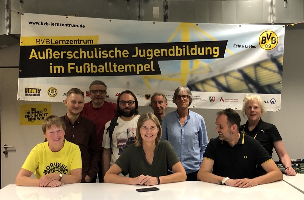 GRÜNE Sommertour - Fußball und Jugendarbeit im BVB-Lernzentrum Auf ihrer Sommertour hat die GRÜNE Ratsfraktion am vergangenen Donnerstag das BVB-Lernzentrum im Westfalenstadion besucht. „Wir wollen mit politischer Jugendarbeit insbesondere bildungsbenachteiligte Jugendliche mit Hilfe des Fußballs für eine offene und vielfältige Gesellschaft begeistern“, erläutern Johannes Böing und Conny Dietz als Mitarbeiter*innen des Lernzentrums den Anspruch der Einrichtung. Träger des Lernzentrum ist das Fan-Projekt Dortmund. In einem Schulungs- und Gruppenraum unterhalb der Südtribüne im Stadion kommen jährlich um die 3000 Schüler*innen zu Workshops zusammen. Dabei werden unterschiedliche Themen wie Zivilcourage, Diskriminierung, Rassismus. Rechtsextremismus, Flucht und Integration, interkulturelles Lernen oder auch Sexismus behandelt. Und auch ökologische Themen werden im inzwischen eigenen Lerngarten im angrenzenden Kleingartenverein behandelt. Der Fußball dient dabei als roter Faden und wird mit den jeweiligen Themen verknüpft. Aktuell arbeitet das Lernzentrum mit fünf Kooperationsschulen enger zusammen, die Workshops stehen aber auch anderen Schulen zur Verfügung. Verknüpft ist die inhaltliche Arbeit mit einer im Anschluss stattfindenden Stadionführung, Britta Gövert, Mitglied der GRÜNEN im Kinder- und Jugendausschuss: „Das ist ein spannender Ansatz von außerschulischer Jugendarbeit. Insbesondere für bildungsbenachteiligte Jugendliche bietet das Lernzentrum die Möglichkeit, sich über einen ganz anderen Zugang mit gesellschaftlichen Themen auseinanderzusetzen. Das Lernzentrum trägt damit zur Weiterentwicklung einer demokratischen und solidarischen Gesellschaft bei.“ Auf dem beigefügten Bild sehen Sie von links nach rechts: Kerstin Feldhoff, Julian Jansen, Michael Stingl, Leander Schreyer, Britta Gövert, Mario Tenge, Stefan Neuhaus (GRÜNE Ratsfraktion/GRÜNE Mitglieder) Johannes Böing, Conny Dietz (BVB Lernzentrum)