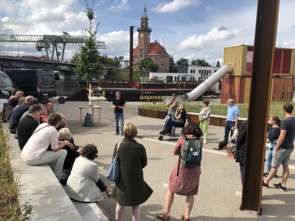 Ausbau des Dortmunder Hafens für die Verschiffung von Schwergut gefordert Der Dortmunder Hafen ist im Umbruch. Im Rahmen ihrer Sommertour unternahm die GRÜNE Ratsfraktion deshalb mit der Dortmunder Hafen AG und der Wirtschafsförderung Dortmund einen Hafenspaziergang, um sich ein Bild vom aktuellen Stand der Entwicklungen zu machen und neue Zukunftsideen zu diskutieren. Quartiersentwicklung an der Speicherstraße „Für uns Grüne war von Anfang an wichtig, dass sich hier ein Quartier für alle entwickelt“, erklärt Svenja Noltemeyer, Ratsmitglied der Grünen im Wirtschaftsförderungsausschuss und Mitglied des Aufsichtsrates der Hafen AG. „Es gibt viele Initiativen, die sich mit Know-how für die Dortmunder Stadtentwicklung - zum Beispiel der des Hafenquartiers - einsetzen. Insbesondere die Kreativwirtschaft beteiligt sich an den planerischen Entwicklungen und konnte Anregungen in die Planungen einbringen. Auch die Speicherstraße nimmt derzeit sichtbar Form an. Dort entsteht ein neues Quartier, das sich mit Lösungen für die Stadt der Zukunft auseinandersetzt. Digitalität wird dabei eine große Rolle spielen.“ Rund 4100 neue Arbeitsplätze und ein Gesamt-Investitionsvolumen von fast 300 Mio. Euro bis 2030: Die Baustellen und erste Fertigstellungen an der Speicherstraße am Rand des Hafens lassen mittlerweile erahnen, wie sich das digitale Quartier in Zukunft neu zeigen wird. Neben dem Fraunhofer Institut werden künftig die Akademie für Theater und Digitalität, der Lensing Media Port für digitale Medienarbeit, das Integrationszentrum Heimathafen und der Innovations- und Gründungscampus im alten Speichergebäude der südlichen Speicherstraße neues Leben einhauchen. Mit Abschluss der Umbauarbeiten werden Promenade und öffentliche Plätze am Hafenbecken am 27. August beim Hafenspaziergang symbolisch der Öffentlichkeit übergeben. Verschiffung von Schwergut ermöglichen „Dortmund hat im Gegensatz zu anderen Städten die Möglichkeit, zu einer ökologisch sinnvollen Transformation des Güterverkehrs beizutragen“, stellt Katrin Lögering, Ratsmitglied der Grünen im Ausschuss für Klimaschutz und ebenfalls Mitglied des Aufsichtsrates der Hafen AG, fest. „Mit der geographisch günstigen Lage, Europas größtem Kanalhafen und der Dortmunder Eisenbahn GmbH haben wir das Potenzial für eine intelligente und ressourcenschonende Transportlogistik von Gütern. Dass das Fraunhofer Institut, das eine jahrzehntelange Erfahrung in der Logistikforschung mitbringt, nun an den Hafen zieht, begrüßen wir GRÜNE ausdrücklich. Es braucht in Zukunft aber auch wieder einen Umschlagplatz für Schwergut, der durch die Neugestaltung der nördlichen Speicherstraße wegfällt. Wir GRÜNE werden dieses Ansinnen des Hafens positiv begleiten, nicht zuletzt, weil Güter in Dortmund immer noch hauptsächlich per LKW transportiert werden – dabei ist der Gütertransport über Schiff und Schiene bis zu 55% günstiger, spart das 4,5-fache an CO2- Emissionen und entlastet den Verkehr; für uns sieht so intelligente Logistik aus.“