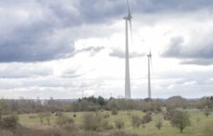Ein weites, vertrocknetes Feld. Hinten zwei Windräder die in den wolkenbehangenen Himmel ragen