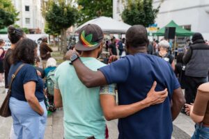 Zwei Männer stehen Arm in Arm in einer Menschenmenge auf dem Kurt-Piehl-Platz und blicken auf das Geschehen. Im Hintergrund beobachten weitere Personen die Veranstaltung.
