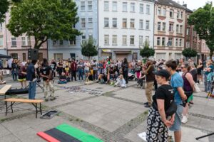 Blick über den Platz und die Zuhörenden während einer Rede. Menschen stehen und sitzen verstreut, vorne auf dem Boden sind zahlreiche Porträtfotos ausgebreitet und eine gerollte Pan-Afrikanische Flagge zu sehen. Wohnhäuser rahmen den Platz ein.