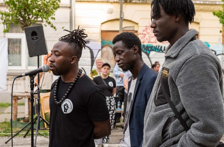 Drei Männer stehen nebeneinander hinter einem Mikrofon auf einer kleinen Bühne beim Gedenken an Mouhamed Lamine Dramé auf dem Kurt-Piehl-Platz. Im Hintergrund Zuhörende und eine mit Graffiti bemalte Hausfassade.