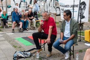 Zwei Männer führen ein Gespräch bei der Diskussionsrunde zum Gedenken an Mouhamed Lamine Dramé. Beide sitzen auf Bänken, einer hält ein Mikrofon.