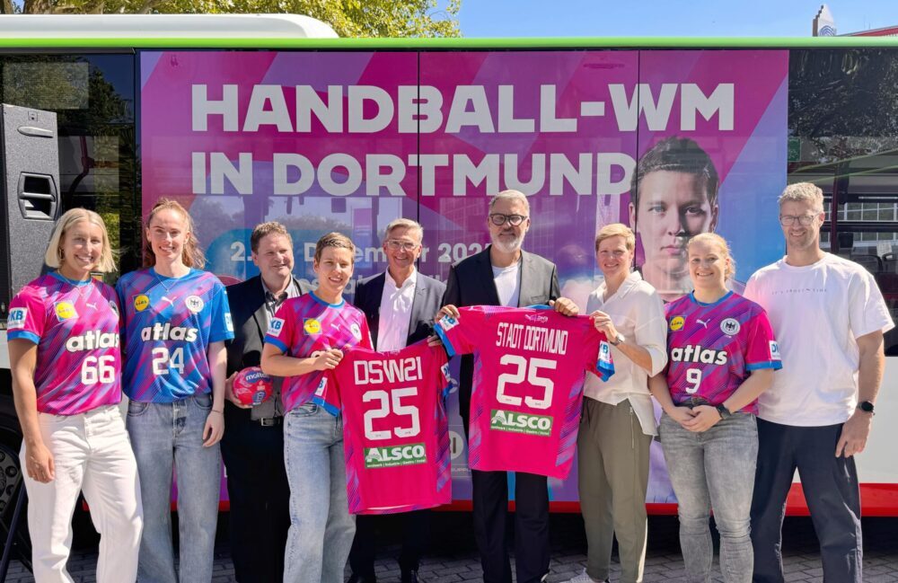 9 Personen stehen vor einem Bus mit der Aufschrift Handball-WM in Dortmund und halten Handball Trikots in die Kamera.