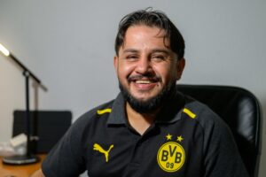 Ein Mann im Borussia Dortmund Polohemd sitzt an einem Schreibtisch. Der erste Knopf des Hemdes ist offen. Er lacht in die Kamera. Sein Haar fällt in einem Mittelscheitel über seine Stirn. Er trägt einen kurzen schwarzen Bart. Im Hintergrund steht eine Schreibtischlampe.