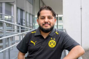 Ein Mann im Borussia Dortmund Polohemd lehnt an einem Geländer. Der erste Knopf des Hemdes ist offen. Er schaut zufrieden.