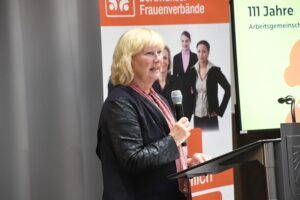 Barbara Wenzel am Mikrofon. Hinter ihr zwei Aufsteller zu der AG Dortmunder Frauenverbände.