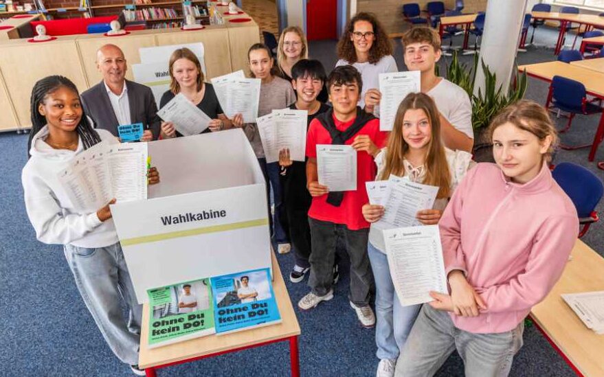 Schulleiter Jörg Skubinn (2.v.l.), die Lehrerinnen Klasina Stache (5.v.l.) und Sylvia Stroh (4.v.r.) freuen sich mit den Schüler:innen vor der Wahlurne