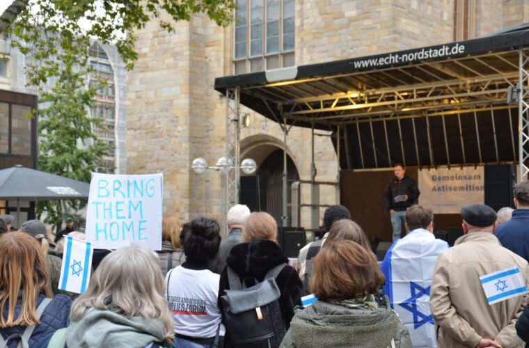 Teilnehmende der Kundgebung sind von hinten zu sehen. Sie blicken auf die Bühne vor der Reinoldikirche, auf der Redebeiträge zum Thema Antisemitismus gehalten werden.