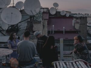 Junge Menschen sitzen auf einem Dach. Die umliegenden Dächer sind mit Sateliten-Schüsseln bespickt. Die Menschen schauen nachdenlich in die Ferne