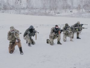 Eine Gruppe an Männern kniet im Schnee. Sie tragen Camouflage-Hosen und Jacken. Sie zielen mit Gewehren auf ein Ziel außerhalb des Bildes.