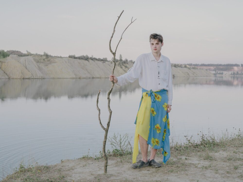Ein Junge Mann mit weissem Hemd und gelb-blauem langen Rock steht in Schlappen vor einem Fluss. Er umgreift einen Stock, der aus dem sandigen Boden ragt. auf dem blauen Teil des Rocks sind gelbe Blumen abgebildet.