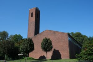 Das Bild zeigt eine braune Kirche. Es ist die ehemalige evangelische Markuskirche an der Gut-Heil-Straße. Bäume stehen davor. Sie ist schlicht und hat einen quadratischen Turm.