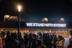 „We stand with Sydney“ steht auf einer Leuchtanzeige und davor die Teilnehmenden der Kundgebung.