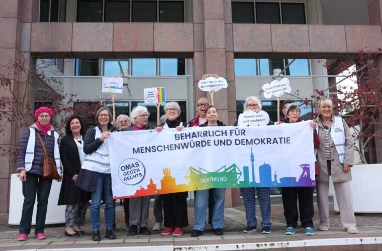 Eine Gruppe von Frauen steht in zwei Reihen vor dem Rathaus und hält Schilder und Transparente in den Händen.﻿