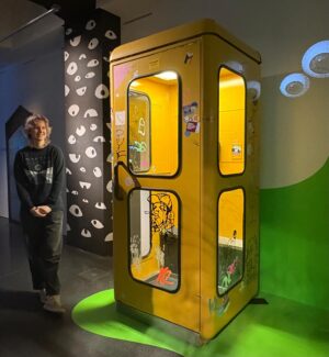 Eine gelbe Telefonzelle mit Künstlerin links daneben stehend.