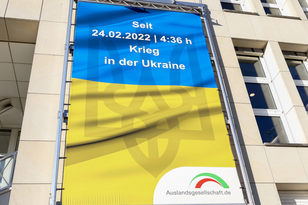 Gelb-blaues Plakat der Auslandsgesellschaft "Seit 24.02.2022, 4:36h Krieg in der Ukraine"