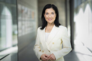 Reiche trägt einen cremefarbenen Blazer und hat die Hände vor dem Bauch zusammengelegt. Sie trägt eine prominente Kreuzkette und lächelt, ohne die Lachmuskeln an den Augen anzuspannen.