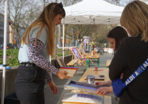 Drei Personen blicken fokussiert auf den Siebdruck auf der Bierbank zwischen ihnen. Die Siebdruckplatte wurde gerade vom Stoff entfernt und man kann einen Blick auf das Ergebnis erhaschen.