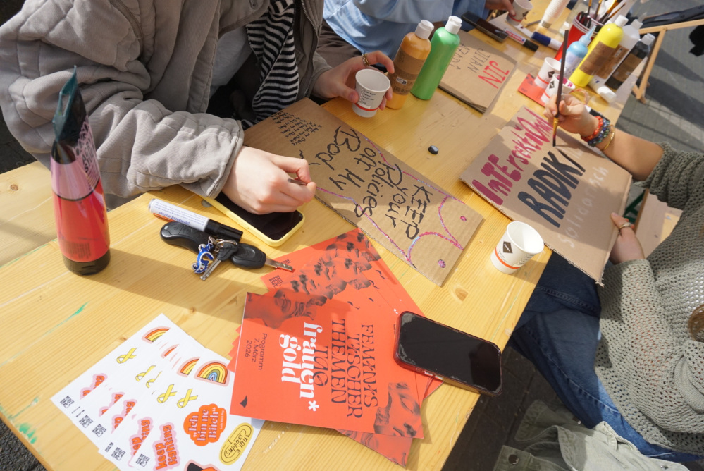 Auf einem Bierbanktisch liegt das Programm des Thementages, feministische Sticker und Plakate von Besucher:innen.