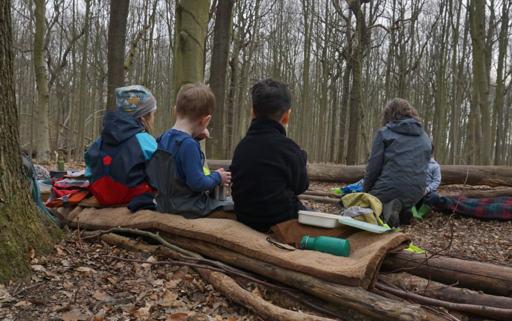 Drei Kinder auf einer Waldbank, mit dem Rück zu sehen. Im Hintergrund eine Erzieherin, die bei einem anderen Kind hockt.