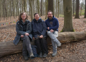 Drei Erwachsene sitzen zur Kamera gewandt auf einem umgekippten Baum. Sie lachen und tragen alle Wanderschuhe. Die Person in der Mitte hält kleine Stöcker in ihrer linken Hand.
