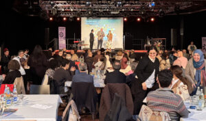 Vor der Bühne des DKH sitzen viele Menschen um Tische und hören dem Interview auf der Bühne zu. Dort stehen zwei Männer und eine Frau.