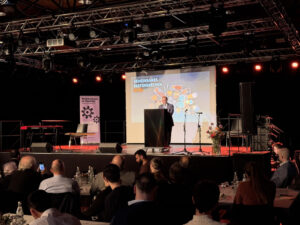 Zu sehen ist eine Bühne. Im Hintergrund ein Banner mit dem Titel "Gemeinsam in Vielfalt" in pastell-lila. Hinter dem Rednerpult wird ein Foto von Essen, das in vielen kleinen Schalen angerichtet ist, gezeigt. Darauf steht: "Gemeinsames Fastenbrechen. Iftar"