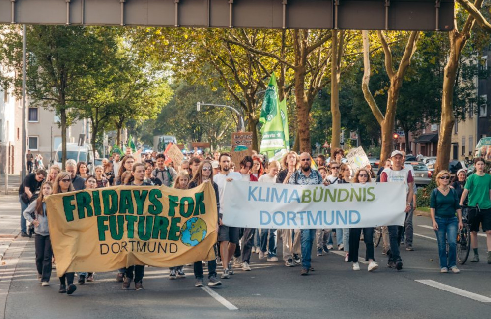 Wie sehen einen Demozug von vorne. Zwei Frontbanner sind zu lesen: "Klima Bündnis Dortmund" und "Fridays For Future Dortmund". Hinter ihnen erkennt man eine Masse von Menschen. Es scheint die Sonne .