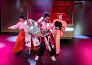 Pinkes Licht. Im Hintergrund ein Bus, der scheinbar aus der Wand herausfährt. Davor die Gurlband in extravaganten Posen und Outfits. Sie performen ein Lied.