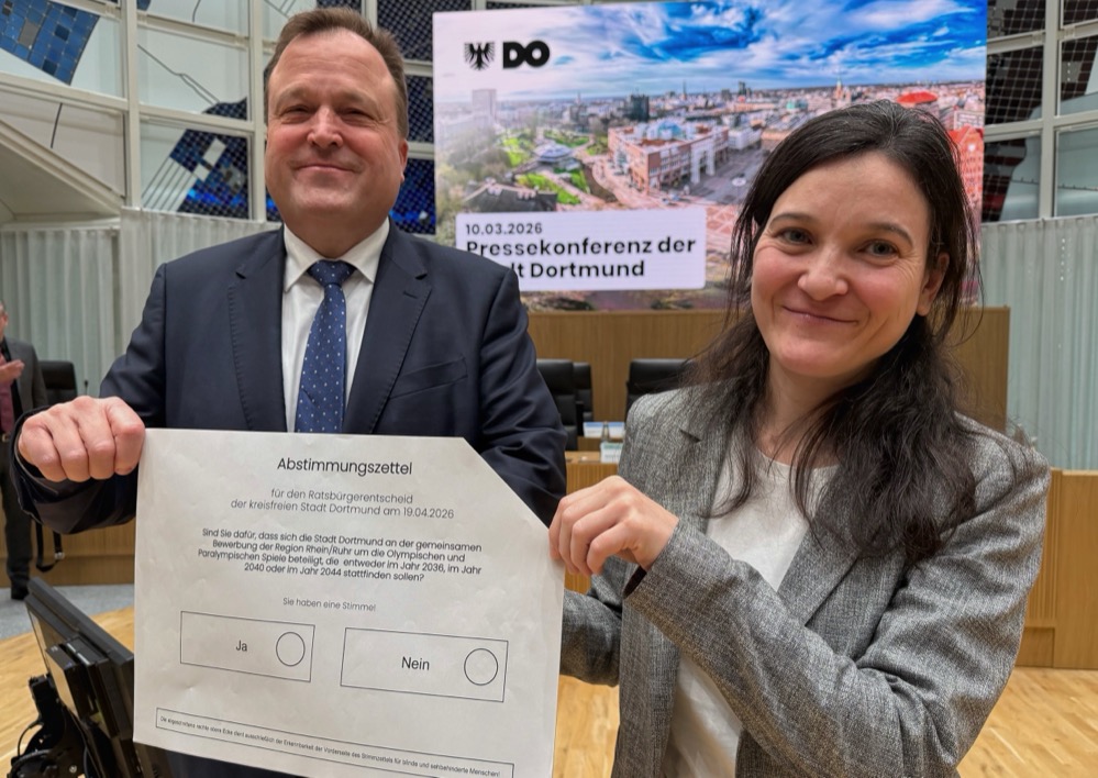 Zwei Personen lächeln in die Kamera. Sie halten einen Abstimmungszettel in den Händen.