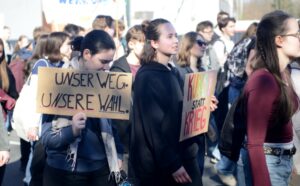 Schülerinnen halten jeweils ein Schild hoch. Auf den Schildern steht: "Unser Weg. Unsere Wahl." und "Kunst statt Krieg"