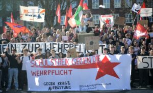 Die streikenden Schüler:innen stehen zum Abschluss auf den Treppen zum Theatervorplatz. Sie halten ihre Banner und Fahnen hoch. Zu sehen ist ein Banner mit der Aufschrift: "Schulstreik gegen die Wehrpflicht"