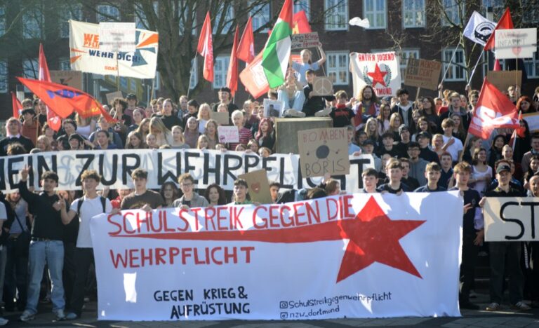 Die streikenden Schüler:innen stehen zum Abschluss auf den Treppen zum Theatervorplatz. Sie halten ihre Banner und Fahnen hoch. Zu sehen ist ein Banner mit der Aufschrift: "Schulstreik gegen die Wehrpflicht"