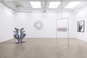 Ein weißer Raum, in dem zwei Skulpturen stehen. Ein Objekt und ein Foto hängen an der Wand