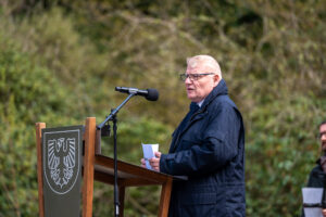 Ein weißer Mann mit weißem Haar und dunkler Jacke an einem Rednerpult mit dem deutschen Adler vorne drauf. Im Hintergurnd Gebüsch.