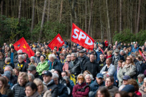 Im Hintergrund ein Wald. Davor Menschen, stehend und sitzend. Keine Sonne. Sie blicken alls nach vorne. Ein paar rote Parteifahnen werden geschwungen, deutlich zu sehen ist die der SPD.