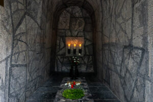 Die Krypta ist grau und mit einem Mosaik versetzt. Ganz hinten leuchten drei Kerzen. Auf dem Boden liegt ein Blumenkranz.