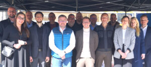 Rund 15 Menschen stehen unter einem Pavillion und lächeln in die Kamera. Ihre Gesichter sind im Shcatten, ihre Körper von der Sonne beschienen.