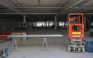 Ein roter Hebelaster und Metallplatten auf Stützen abgelegt im Vordergrund. Im Hintergrund eine weite Halle, ganh hinten: kleine Fenster. Alles wirkt grau.