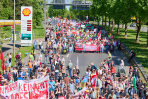 Große Demonstration mit vielen Menschen und Bannern auf einer breiten Straße in Dortmund am ersten Mai, im Hintergrund eine Shell-Tankstelle und grüne Bäume entlang der Straße