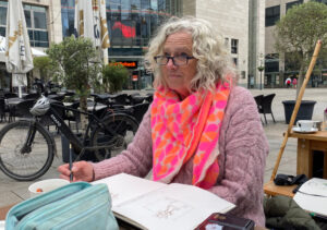 Eine Frau mit grauen, langen Haaren und einer Brille. Ein pink-rosanes Outfit.